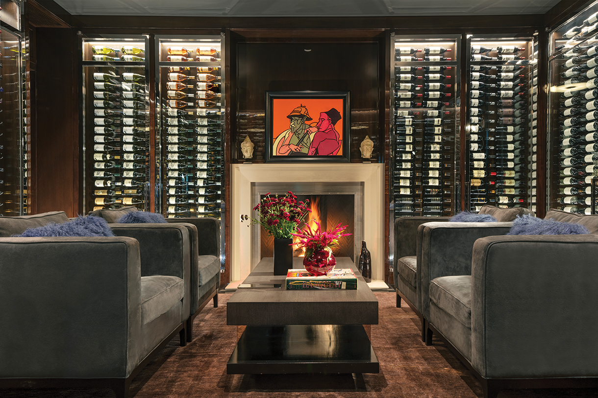 A fully stocked wine library with walnut casework and walls furnished and decorated with charcoal-colored armchairs, modern coffee table, limestone fireplace and an Italian abstract painting displayed on the mantle. 